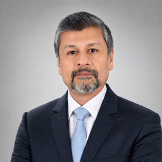 Professional head-and-shoulders portrait of a man in a navy suit and light blue tie against a neutral grey background.