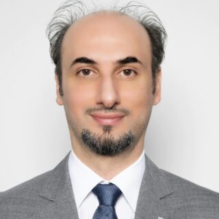 Professional head-and-shoulders portrait of a man in a gray suit and blue tie, light background, slight smile.