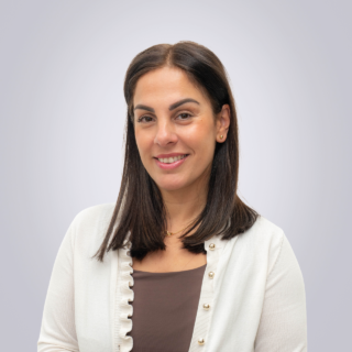 Portrait of a smiling woman with dark hair, wearing a beige cardigan over a brown top, against a light gray background.