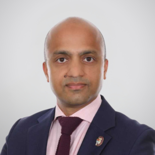Professional headshot of a bald man in a navy suit, pink shirt, and maroon tie against a gray gradient background