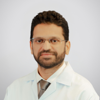 Portrait of a male doctor wearing glasses, a white lab coat, and a tie, facing the camera against a neutral grey background.