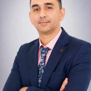 Man in a navy blazer, pink shirt, and floral tie poses with arms crossed against a gray background.